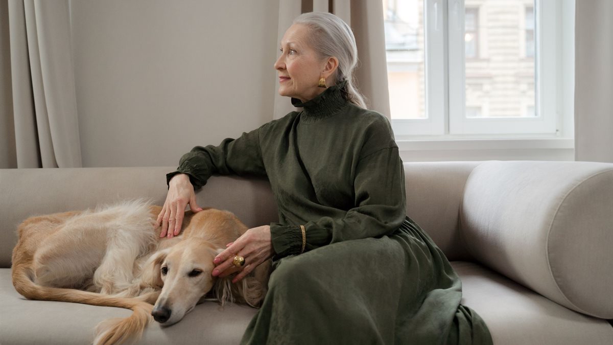 An older woman sitting on a sofa with her greyhound resting beside her