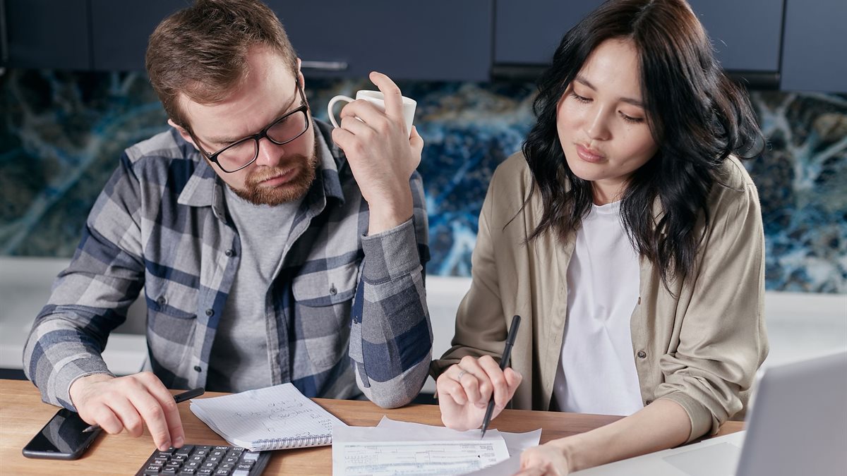 A couple at a kitchen table reviewing household bills with a calculator and laptop